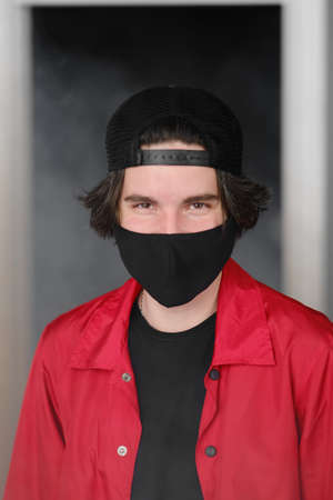 Portrait of a young man of 25-30 years old in a black protective mask, cap and red jacket. A man posing on a gray background in the smoke. Coronavirus protection.の写真素材
