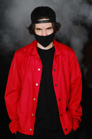 Portrait of a young man of 25-30 years old in a black protective mask, cap and red jacket. A man posing on a gray background in the smoke. Coronavirus protection.の写真素材