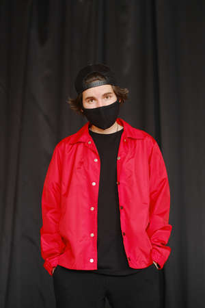 Portrait of a young man 25-30 years old in a black protective mask, black cap and red jacket. Young fashionable man posing over bright background. Fashion photography conceptの写真素材
