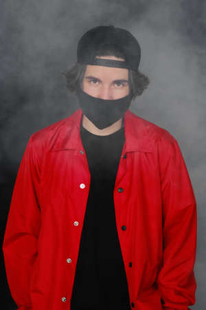 Portrait of a young man of 25-30 years old in a black protective mask, cap and red jacket. A man posing on a gray background in the smoke. Coronavirus protection.の写真素材