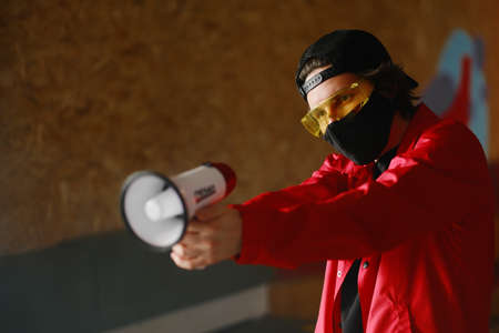 A young man of 25-30 years old in a black protective mask, yellow goggles, a cap and a red jacket holds a megaphone against a dark background. Protest conceptの写真素材