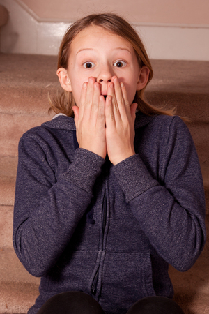 Young teenage girl in gray jacket with her hands covering her mouth to express shock, horror and surprise, indoor background.の写真素材