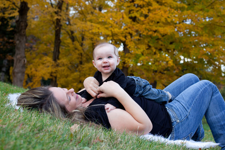 Happy mother playing with her baby girl on some grass with autumn trees in the background.の写真素材