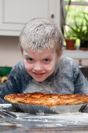 boy covered in flour by pie / A smiling boy covered in flour and a baked pie on a table in front of him.の写真素材