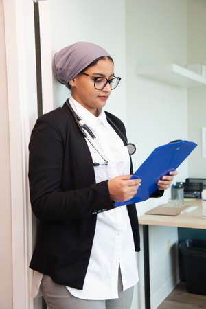 a doctor wearing a hijab and stethascope looks at a medical chart in her office