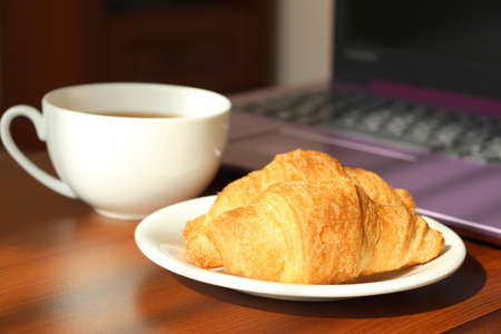 White cup with black tea and croissants in a saucer on the work table. Remote work at home. Snack...
