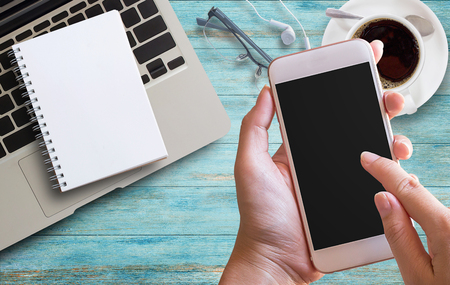 Smart phone in female hand with office desk with laptop, coffee and glasses on wooden table background. Workplace. Top view with copy space. Learning Education. Knowledge and Training E-Learning Skills Start Up.の写真素材