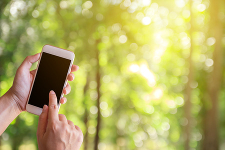 Women hand shows mobile smartphone in vertical position with blurred green bokeh background and sunlight use for mockup template. Green bokeh abstract background for design or graphic display montage.の写真素材