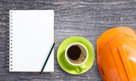 Top view with orange safety helmet, blank notepad and pencil with cup of coffee on office wood table...