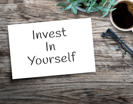 Text sign showing Invest In Yourself. on wood table with flower,glasses and cup of coffee on wood...