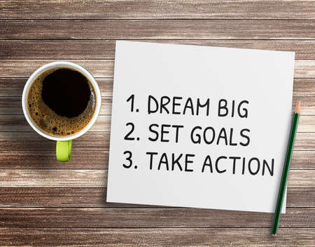 Inspirational handwriting on a white paper with a cup of coffee and pencil on wood table background. text with Dream big, set goals, take actionの写真素材