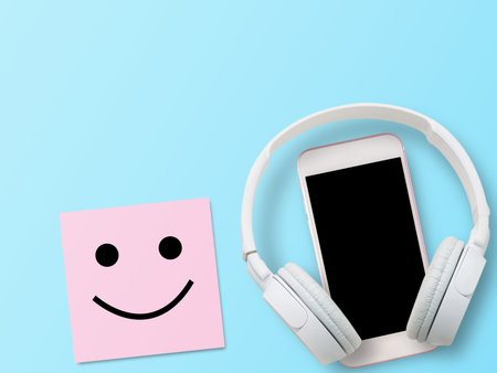 White headphone, smartphone and smiley symbol in pink sticky note on blue background. Happiness and...