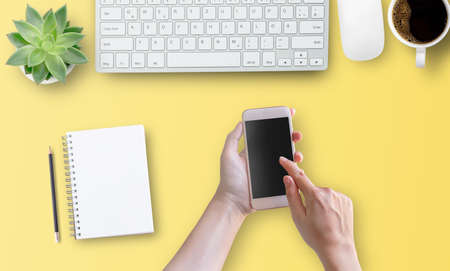 Top view with woman hand using mobile phone with empty screen on yellow table background. Office desktop and business marketing concept.の写真素材