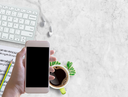 Top view hand holding smartphone,white earphone,pencil,cup of coffee and sheet music on white marble background.の写真素材