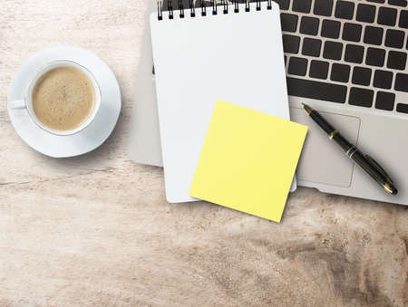 Empty yellow adhesive paper, notebook on laptop keyboard, pen and coffee cup on office desk background. Top view with copy space.の写真素材