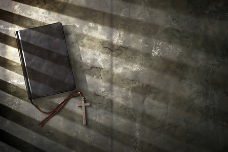 A dark leather notebook lies on a cracked concrete surface, accompanied by a wooden cross. Dramatic shadows enhance the composition, evoking themes of faith and contemplation.の写真素材