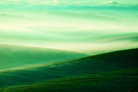 This serene image captures gentle green hills enveloped in mist at dawn, creating a tranquil and dreamy atmosphere, perfect for nature lovers and peaceful landscapes.の写真素材