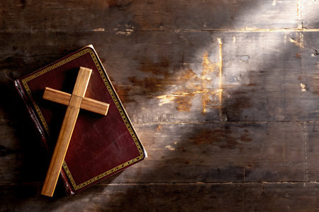 A wooden cross lies elegantly on an open Bible, creating a serene atmosphere on a dark wooden table. The interplay of light and shadow evokes a sense of spirituality and reflection.の写真素材