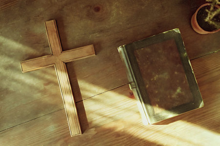 A beautiful wooden cross lies next to a leather bound book on a rustic wood surface with soft...