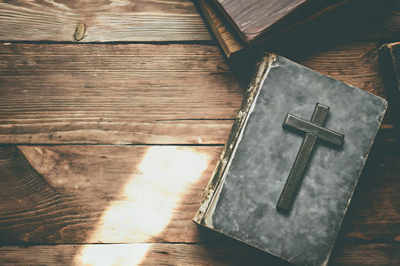 An old book with a cross on its cover rests on a wooden table, illuminated by soft natural light,...