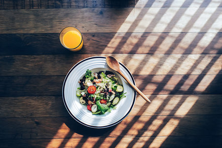 A colorful salad with fresh vegetables and a glass of orange juice rests on a wooden table, illuminated by sunlight shadows, perfect for a healthy meal.の写真素材