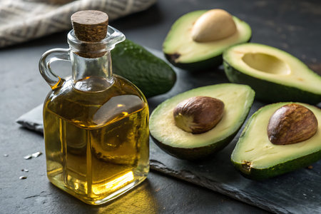 A glass bottle filled with fresh avocado oil stands beside halved avocados on a dark slate surface,...
