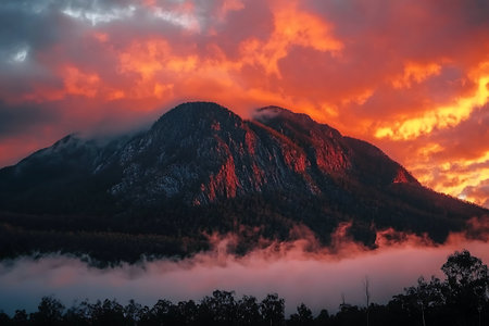 A striking sunset casts vibrant orange and red hues over a rugged mountain peak, enveloped in wispy clouds and mist. The serene atmosphere captivates nature lovers and adventurers alike.の写真素材