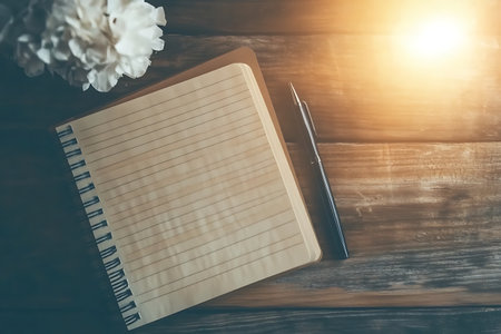 A blank notebook with a pen and a delicate flower rests on a rustic wooden table, illuminated by soft morning light, offering a serene atmosphere for writing.の写真素材