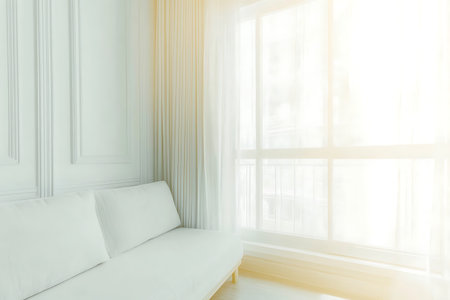 A bright and minimalist living room features a white sofa and sheer curtains, creating a serene ambiance enhanced by natural sunlight streaming through the window.の写真素材