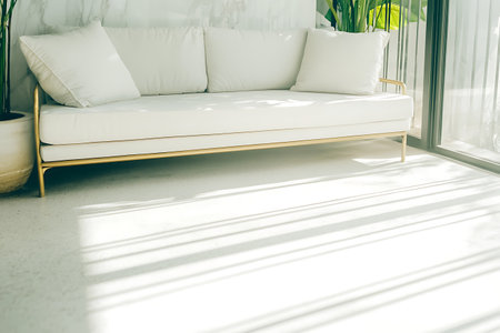 A modern minimalist white sofa with gold legs sits elegantly in a sunlit living room, surrounded by greenery. The bright space radiates comfort and style.の写真素材