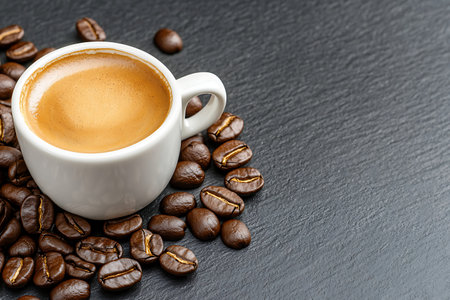 A close-up image of a creamy espresso in a white cup, surrounded by rich coffee beans on a dark slate surface, perfect for food and drink themes.の写真素材