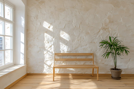 A serene interior scene showcasing a simple wooden bench and a green potted plant beside a sunlit wall. The bright atmosphere invites tranquility and freshness.の写真素材