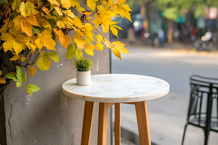 A round marble table with wooden legs nestled beside vibrant yellow leaves captures the essence of...