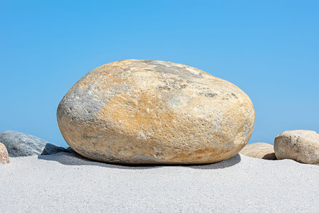 A large smooth stone rests on sandy shore, contrasting beautifully with a clear blue sky, showcasing...