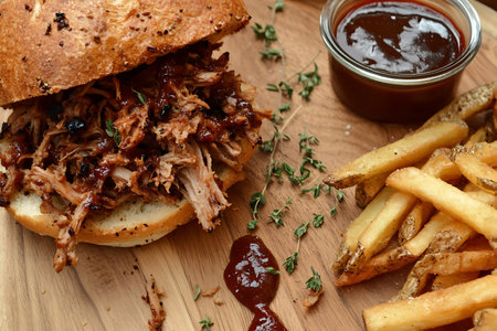 This image features a pulled pork sandwich generously topped with barbecue sauce, served alongside crispy fries and fresh herbs, perfect for any meal.の写真素材