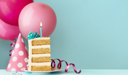 Slice of birthday cake with blue birthday candle, party hat, streamers and birthday balloons ready for a birthday partyの写真素材