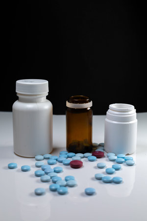 two plastic bottles and one glass with colored pills on a white table with a black background....