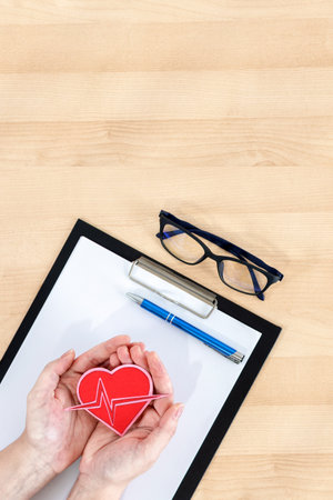 Female hands holding heart, clipboard and glasses on wooden desk. top view.の写真素材