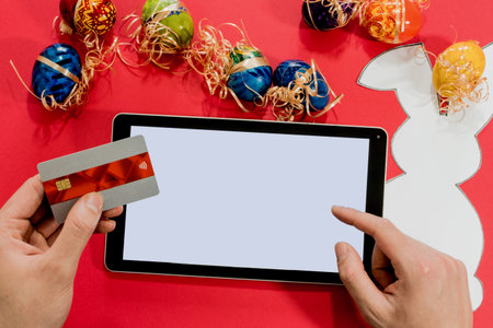 Top view of male hands with tablet, credit card and easter eggs, paper rabbit. On red background....