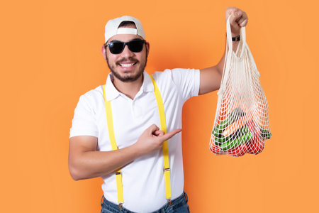 Hipster man holding eco friendly shopping bag with vegetables, posing isolated on orange background in studio.の写真素材