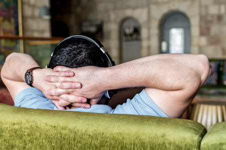 Rear view of a man relaxing sitting on a couch with the hands on the head, in living room at home.
