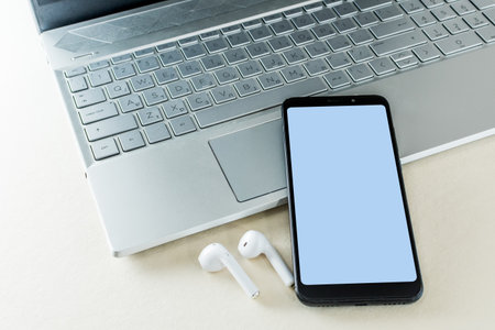 Smart phone with white screen and earphone next laptop, on top of white table workspace. Mockup of modern black color Smart phone.の写真素材