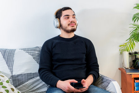 Front view of a happy man with headphones using a mobile phone on line sitting on a couch in the living room at home.の写真素材
