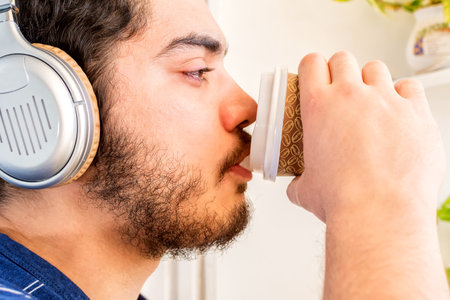 Young man with beard looking out and listening to music using wireless headphones drinking coffee, on the balcony.の写真素材