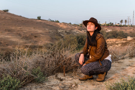 Young woman in hat and brown leather jacket, looking into the distance, squatting in natural park. Travel, lifestyle and tourism concept.の写真素材