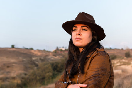 Young woman in hat and brown leather jacket, looking into the distance, in natural park. Travel, lifestyle and tourism concept.の写真素材