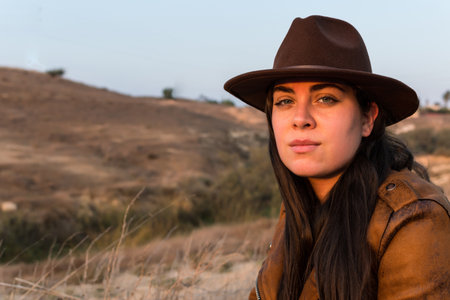 Young woman in hat and brown leather jacket, looking at camera, in natural park. Travel, lifestyle and tourism concept.の写真素材