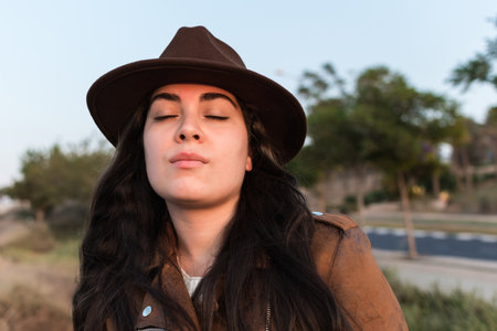 Young woman in hat and brown leather jacket, with closed eyes, in natural park. Travel, lifestyle and tourism concept.の写真素材