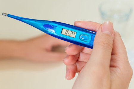 Female hands holding a digital thermometer, tablets in blister packs, a glass of water, on desk....
