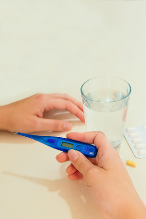 Female hands holding a digital thermometer, tablets in blister packs, a glass of water, on desk. Concept of fever, flu or virus.の写真素材